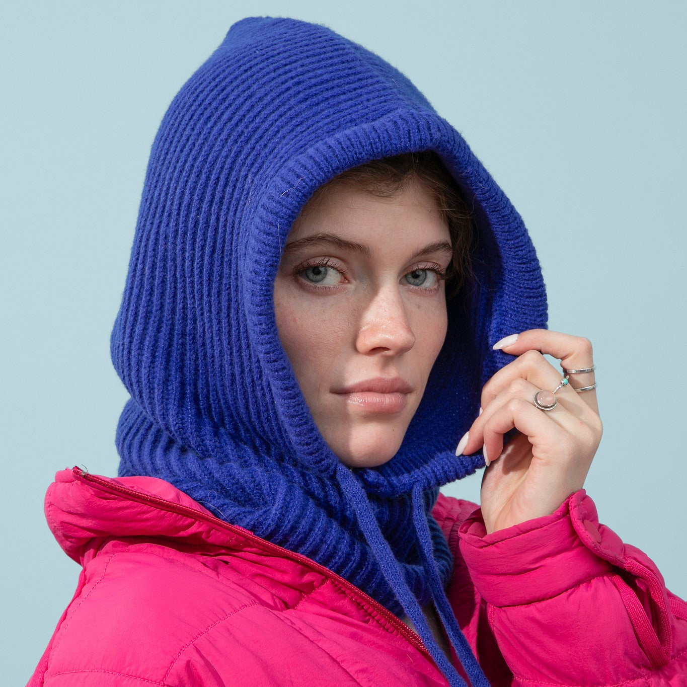 Person wearing a bright pink puffer jacket with a blue knitted hood against a light blue background