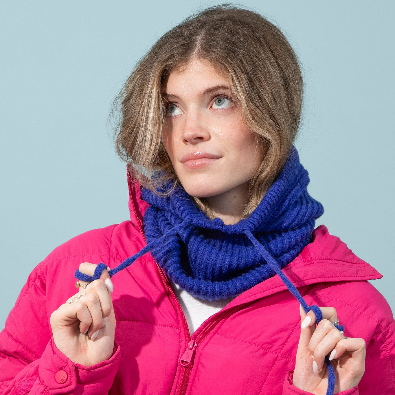Woman wearing a bright pink jacket and blue knitted cowl against a light blue background