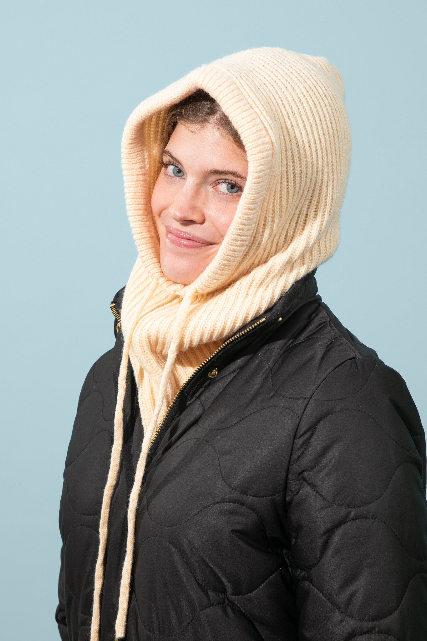 Person wearing a black coat and beige knitted hood against a light blue background